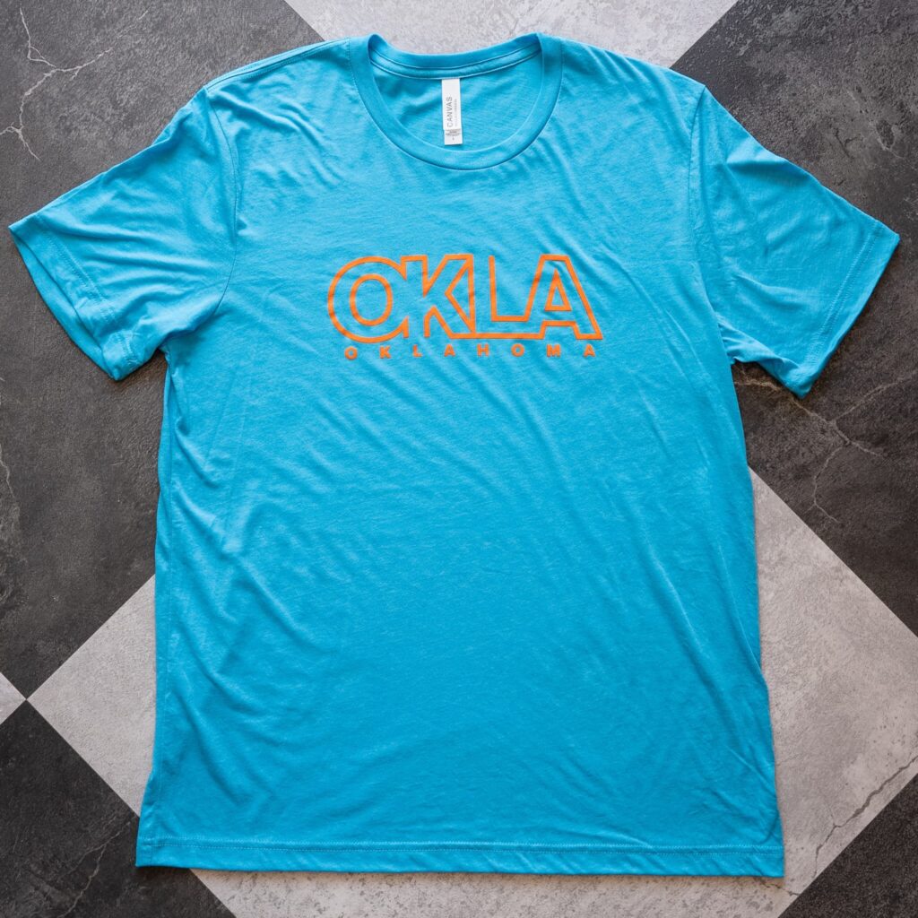 Unisex blue short sleeve tee with "OKLA" in orange block lettering on front for men's or women's style or souvenir gift.