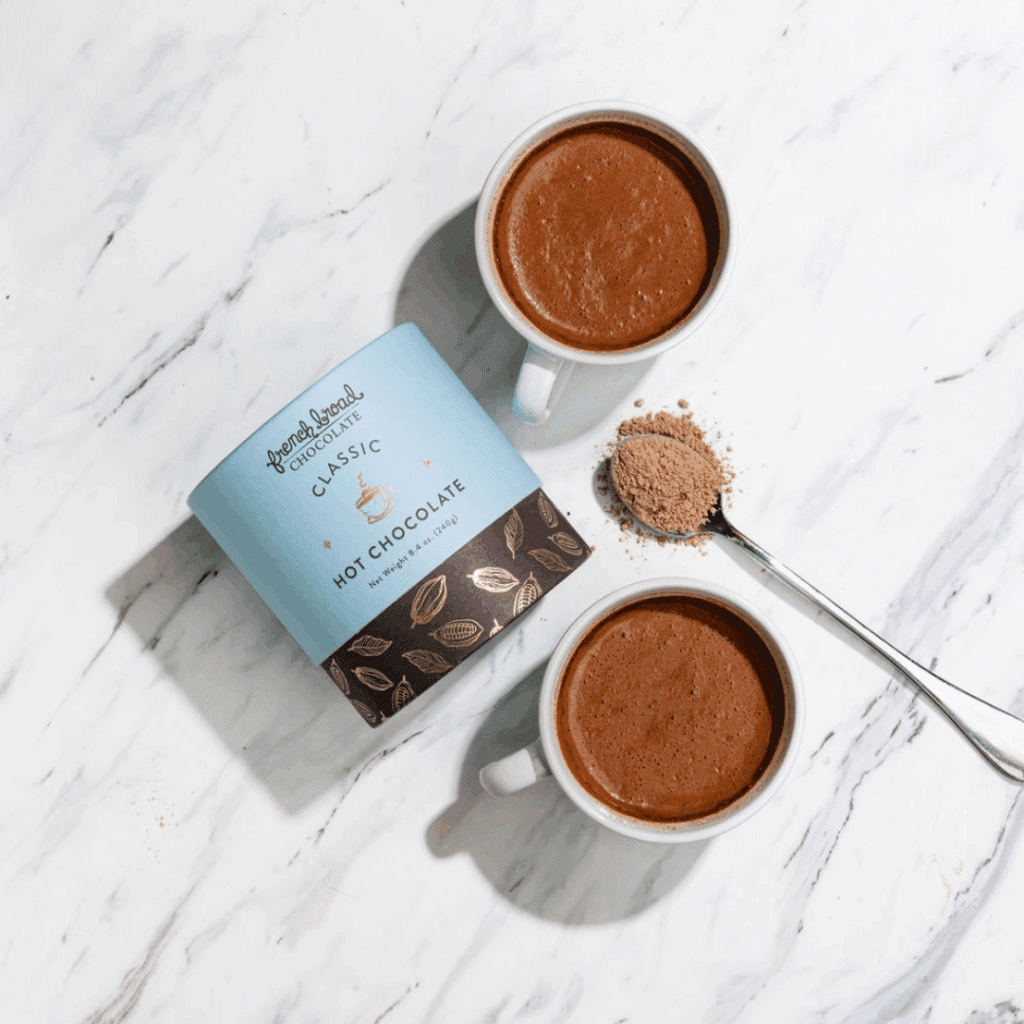 French Broad Dark Chocolate hot chocolate mix in canister next to two prepared mugs of cocoa mix and a spoonful of dry mix