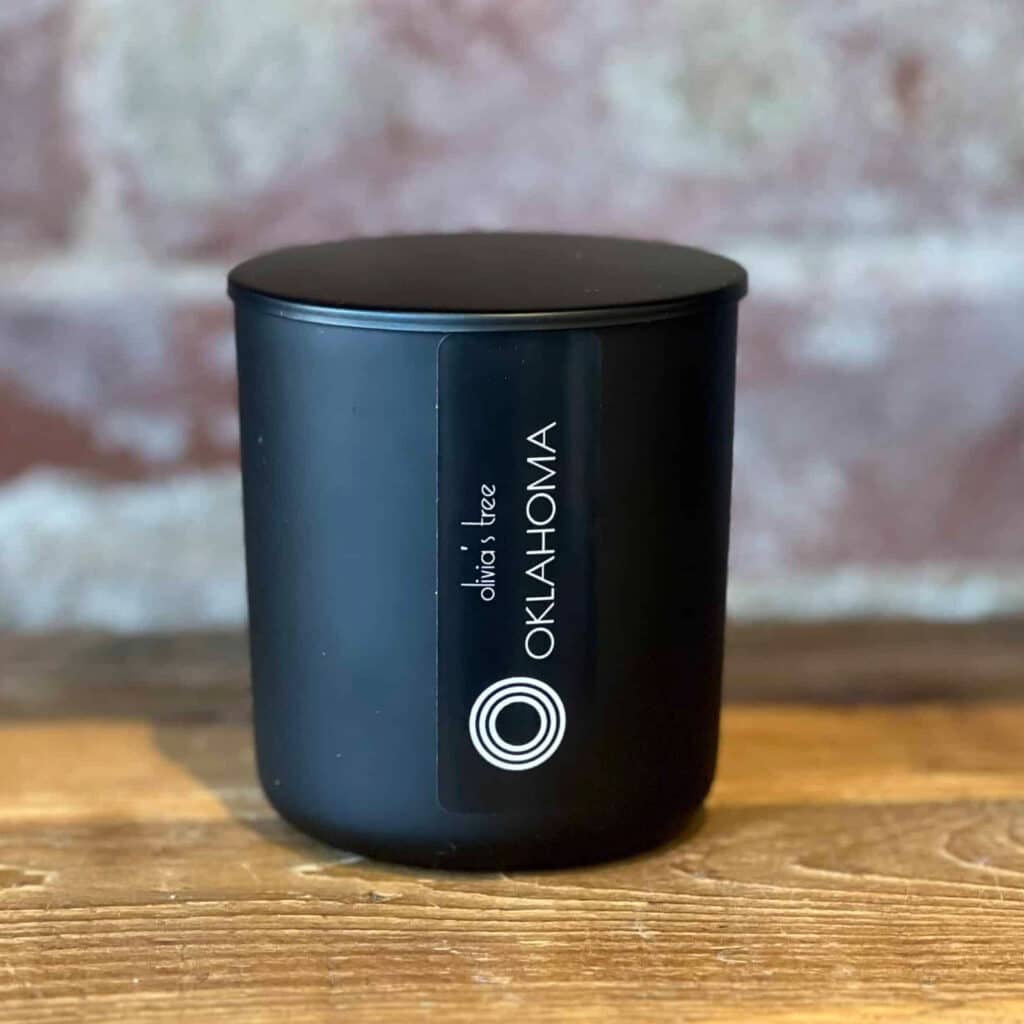 Matte black candle vessel with a matching lid and minimalist white logothat says Oklahoma, displayed on a wooden surface against a softly blurred brick background.