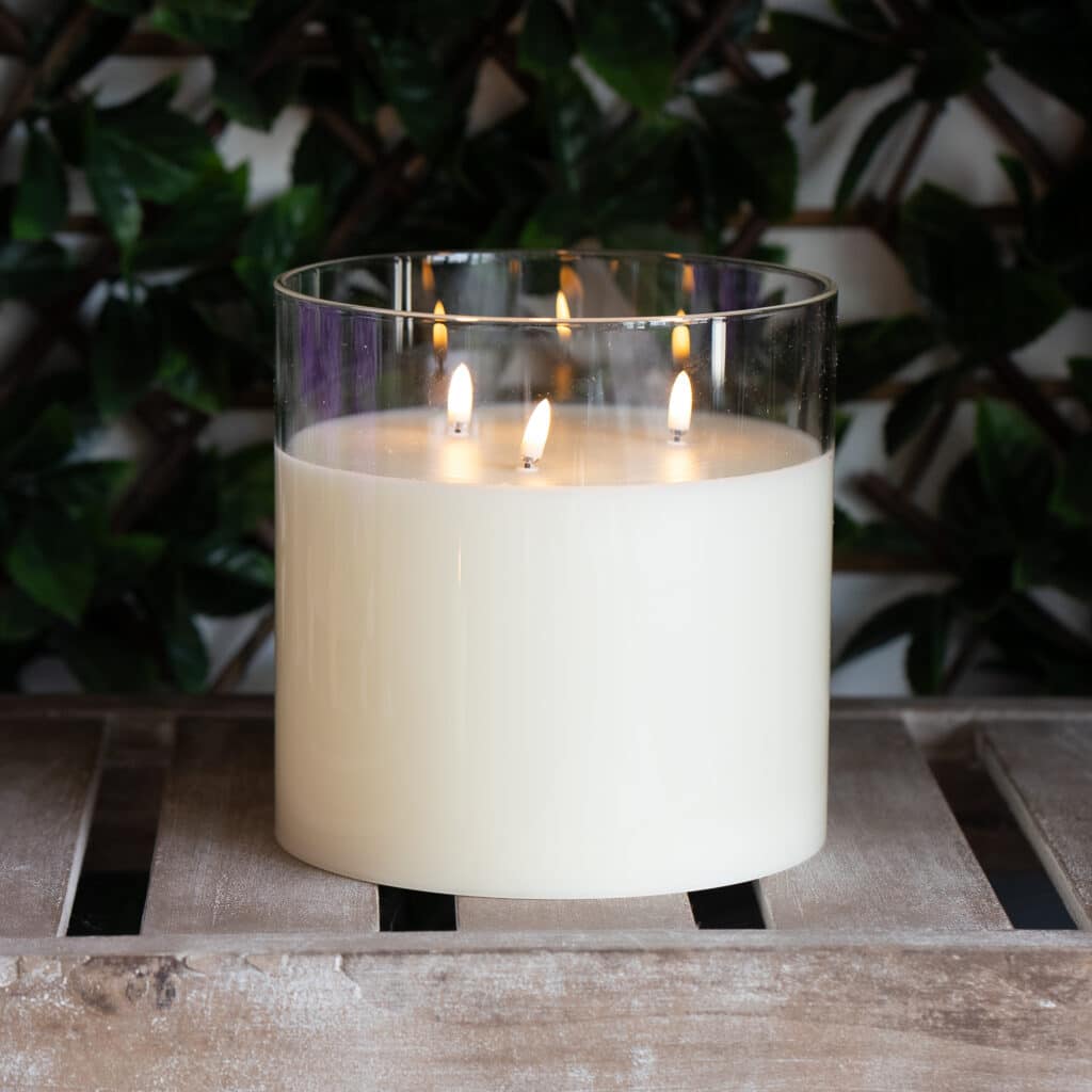 Three-wick battery operated white candle in a clear glass vessel, glowing softly on a tiled tabletop with leafy greenery blurred in the background. The minimalist candle design creates a warm, elegant ambiance.